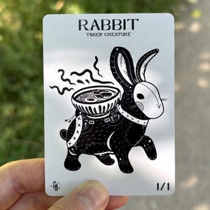 May include: A white rectangular game card featuring a black and white illustration of a rabbit wearing a mask and carrying a bowl of steaming ramen on its back. The text RABBIT TOKEN CREATURE is printed at the top, with 1/1 in the bottom right corner.