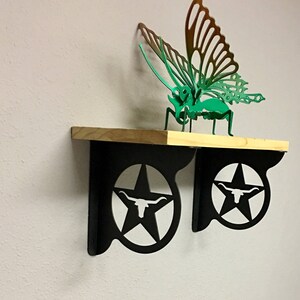 May include: A wooden shelf with two black metal brackets. The brackets feature a star with a longhorn steer inside. A green metal butterfly is sitting on the shelf.