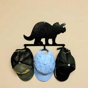 Puede incluir: Un gancho de pared de metal negro con silueta de dinosaurio con tres sombreros colgando de él. Los sombreros son una gorra de malla negra, una gorra de béisbol azul claro y una gorra de béisbol negra.