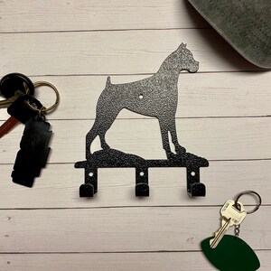 May include: Black metal key holder shaped like a boxer dog with three hooks. The key holder is on a white wooden background.