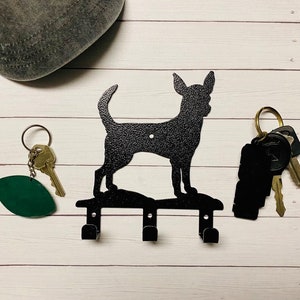 Puede incluir: Porta llaves de metal negro con forma de silueta de perro Chihuahua con tres ganchos. El porta llaves est&aacute; montado en una superficie de madera blanca.