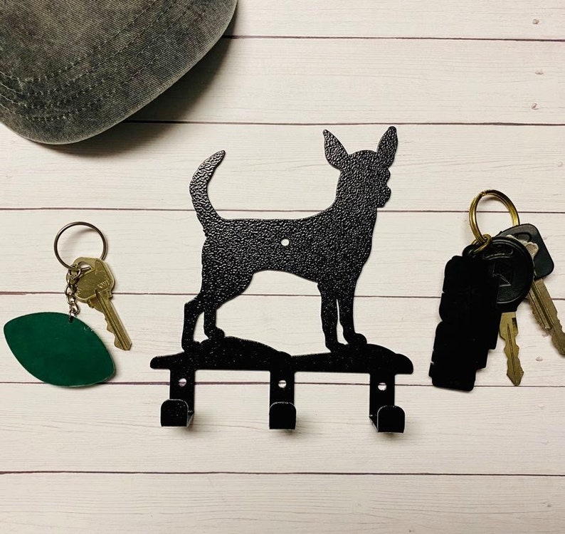 Puede incluir: Porta llaves de metal negro con forma de silueta de perro Chihuahua con tres ganchos. El porta llaves est&aacute; montado en una superficie de madera blanca.