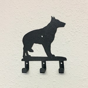 Puede incluir: Llavero de metal negro montado en la pared con la silueta de un perro pastor alem&aacute;n. El llavero tiene tres ganchos debajo de la silueta del perro, perfectos para colgar llaves u objetos peque&ntilde;os. El perro est&aacute; de pie.