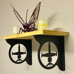 Puede incluir: Una estantería de madera con soportes metálicos negros con la silueta de un avión. La estantería sostiene una escultura de mariposa morada y dos pequeños candelabros blancos. Los soportes están fijados a una pared blanca.