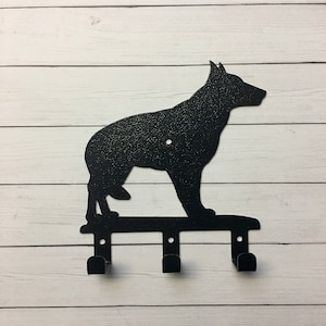 Puede incluir: Un llavero de metal negro con la silueta de un perro pastor alem&aacute;n. El llavero tiene tres ganchos debajo de la forma del perro, perfectos para colgar llaves u objetos peque&ntilde;os. El perro est&aacute; de pie sobre una barra horizontal.