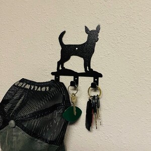 Puede incluir: Porta llaves de metal negro con forma de silueta de chihuahua con tres ganchos. Una gorra de b&eacute;isbol negra y gris cuelga de un gancho y un juego de llaves cuelga de otro.