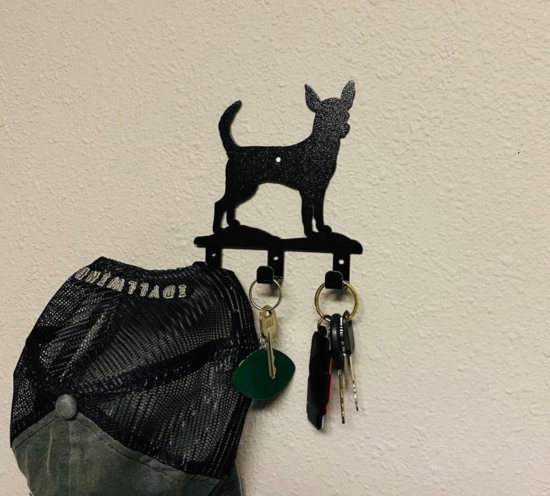 Puede incluir: Porta llaves de metal negro con forma de silueta de chihuahua con tres ganchos. Una gorra de b&eacute;isbol negra y gris cuelga de un gancho y un juego de llaves cuelga de otro.
