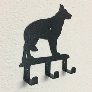 Puede incluir: Un perchero de pared de metal negro con la silueta de un perro pastor alem&aacute;n sobre tres ganchos individuales. El metal tiene un acabado texturizado e incluye orificios pretaladrados para montar la pieza en una superficie plana.