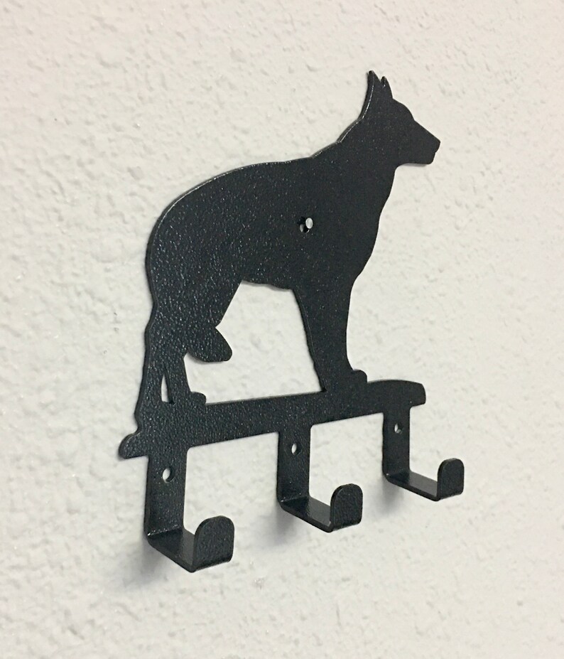 Puede incluir: Un perchero de pared de metal negro con la silueta de un perro pastor alem&aacute;n sobre tres ganchos individuales. El metal tiene un acabado texturizado e incluye orificios pretaladrados para montar la pieza en una superficie plana.