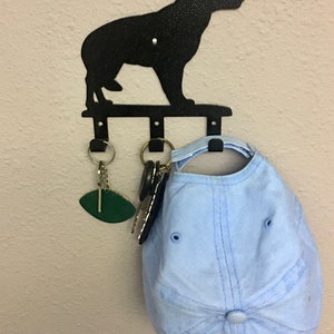 Puede incluir: Llavero de metal negro con forma de perro con tres ganchos. Una gorra de b&eacute;isbol azul claro y llaves cuelgan de los ganchos. El llavero est&aacute; montado en una pared beige.