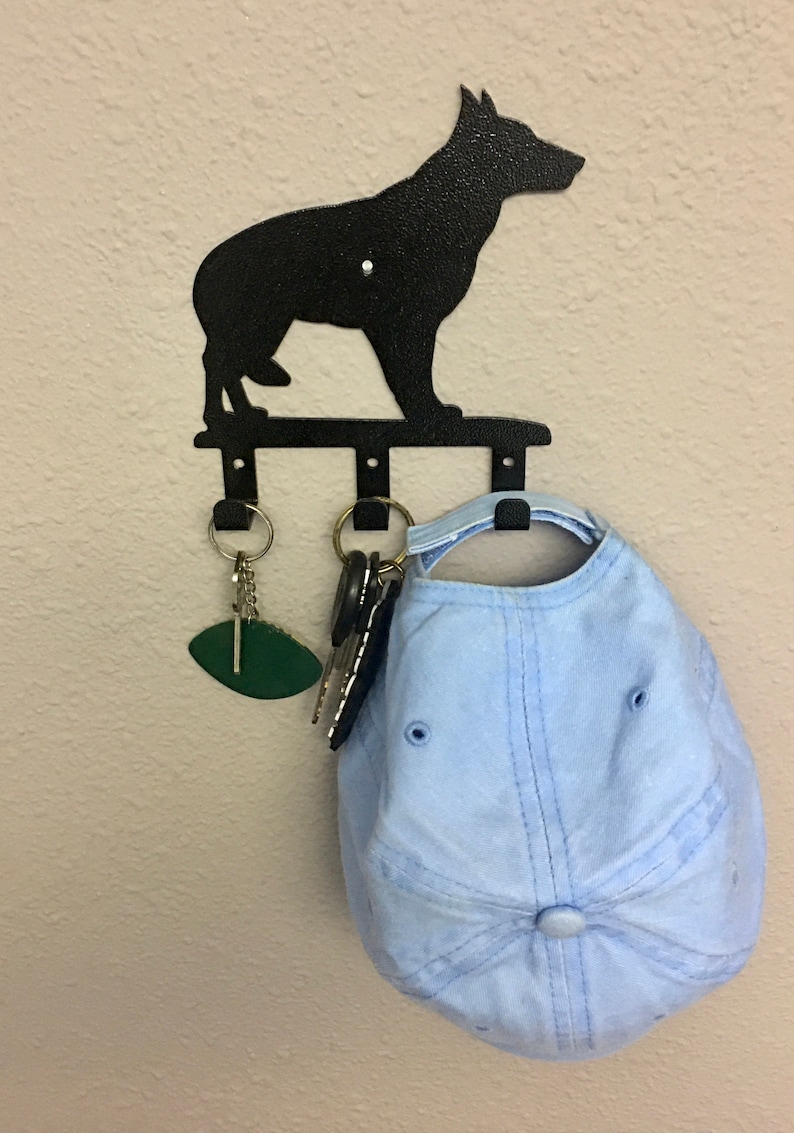 Puede incluir: Llavero de metal negro con forma de perro con tres ganchos. Una gorra de b&eacute;isbol azul claro y llaves cuelgan de los ganchos. El llavero est&aacute; montado en una pared beige.