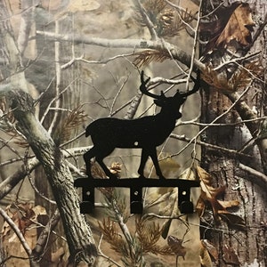 Op de afbeelding: Zwarte metalen sleutelhanger met hert-silhouet en drie haken. Het hert staat op een metalen balk met een camouflage achtergrond.