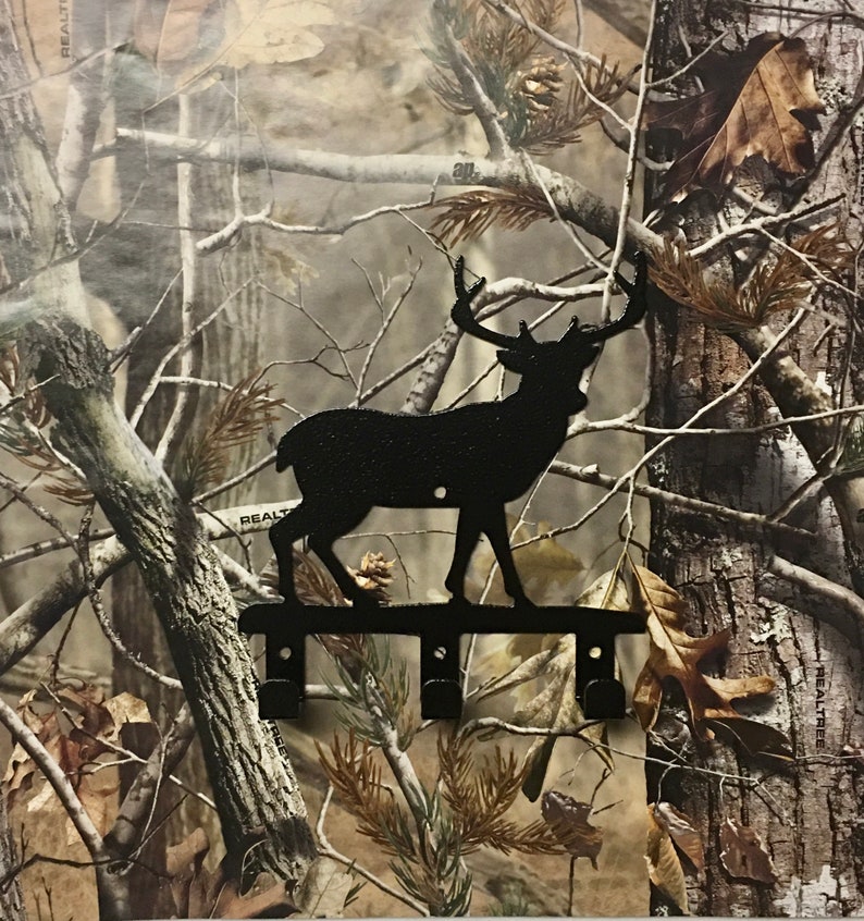 Op de afbeelding: Zwarte metalen sleutelhanger met hert-silhouet en drie haken. Het hert staat op een metalen balk met een camouflage achtergrond.