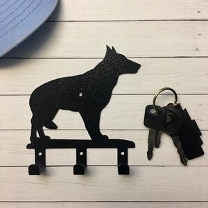Puede incluir: Llavero de metal negro con la silueta de un perro, con tres ganchos para llaves. El llavero est&aacute; montado sobre una superficie de madera blanca. Un juego de llaves cuelga de los ganchos. Una gorra de b&eacute;isbol azul claro est&aacute; en el fondo.