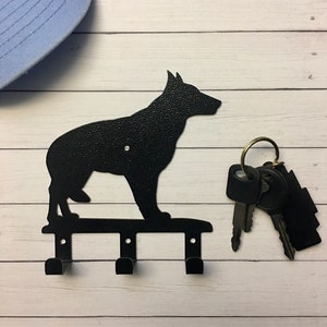 Puede incluir: Llavero de metal negro con la silueta de un perro, con tres ganchos para llaves. El llavero est&aacute; montado sobre una superficie de madera blanca. Un juego de llaves cuelga del llavero. Una gorra de b&eacute;isbol azul claro est&aacute; en el fondo.