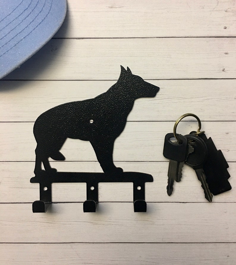 Puede incluir: Llavero de metal negro con la silueta de un perro, con tres ganchos para llaves. El llavero est&aacute; montado sobre una superficie de madera blanca. Un juego de llaves cuelga del llavero. Una gorra de b&eacute;isbol azul claro est&aacute; en el fondo.