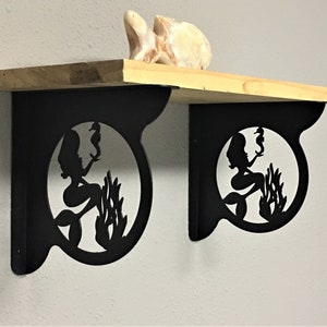 May include: Two black metal shelf brackets with a mermaid silhouette design. Each bracket features a mermaid holding a seahorse in a circular frame with seaweed.