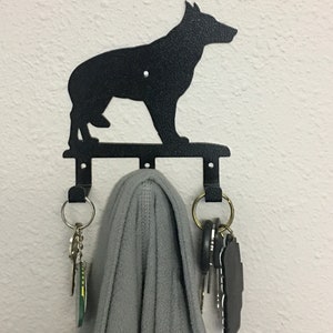 Puede incluir: Llavero de metal negro con la silueta de un perro pastor alem&aacute;n. El llavero tiene tres ganchos y est&aacute; montado en una pared blanca. Una bufanda gris y llaves cuelgan de los ganchos.