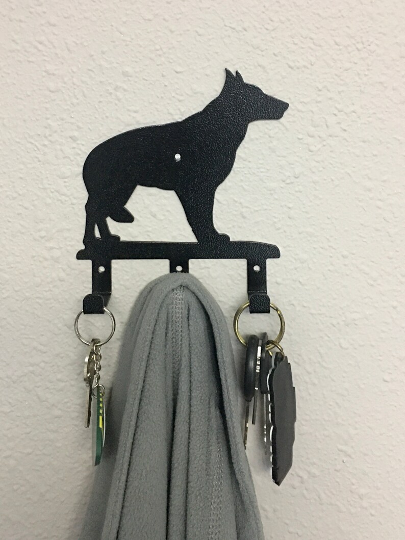 Puede incluir: Llavero de metal negro con la silueta de un perro pastor alem&aacute;n. El llavero tiene tres ganchos y est&aacute; montado en una pared blanca. Una bufanda gris y llaves cuelgan de los ganchos.