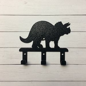 May include: Black metal dinosaur key holder with three hooks. The dinosaur is a triceratops with a textured finish.