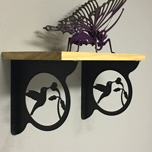 Puede incluir: Dos soportes de estante de metal negro con un diseño de silueta de colibrí. Cada soporte presenta un recorte circular con un colibrí bebiendo néctar de una flor. Los soportes están unidos a un estante de madera.