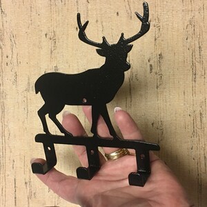Op de afbeelding: Zwarte metalen hert silhouet wandhaak met drie haken. Het hert staat op een metalen basis.