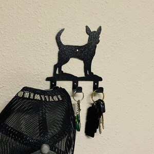Puede incluir: Porta llaves de metal negro con forma de silueta de chihuahua con tres ganchos. Una gorra de b&eacute;isbol de malla negra cuelga de un gancho y un juego de llaves cuelga de otro gancho.