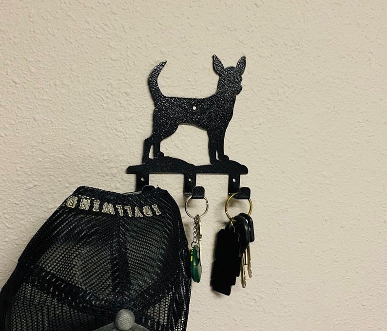 Puede incluir: Porta llaves de metal negro con forma de silueta de chihuahua con tres ganchos. Una gorra de b&eacute;isbol de malla negra cuelga de un gancho y un juego de llaves cuelga de otro gancho.