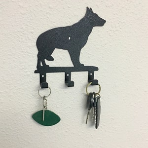 Puede incluir: Llavero de metal negro con la silueta de un perro. El llavero tiene tres ganchos y est&aacute; montado en una pared blanca. Una etiqueta de llave ovalada verde y un juego de llaves cuelgan de los ganchos.