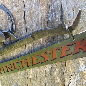 Può includere: Un cartello in metallo con la parola "WINCHESTER" in lettere rosse su sfondo verde. Il cartello è attaccato a un ferro di cavallo e a un gancio di metallo.
