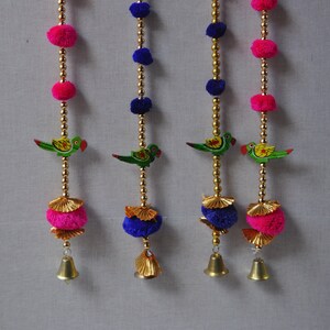 Könnte beinhalten: Drei dekorative Türhänger mit goldenen Ketten, rosa und blauen Pompons, grün bemalten Vögeln und goldenen Glöckchen am unteren Ende.
