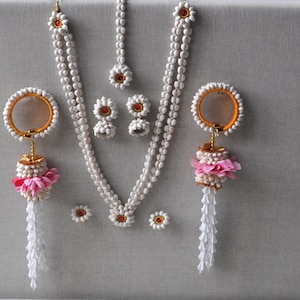 Könnte beinhalten: Ein weißes Blumenketten-Set mit passenden Ohrringen und einem Tikka. Das Set besteht aus weißen Blumen mit goldenen Akzenten und rosa Blumenakzenten an den Ohrringen.