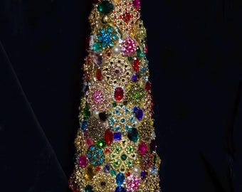 Puede incluir: Una escultura decorativa en forma de cono cubierta con broches de metal dorado, pedrería facetada de colores rojo, azul, verde y rosa, y perlas blancas. La base está rodeada por una banda de pequeños cristales transparentes y perlas.