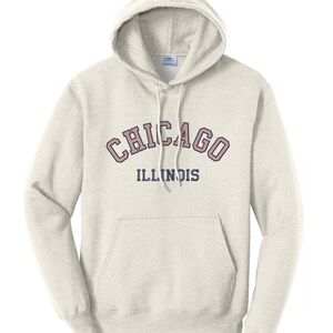 May include: A light beige hooded sweatshirt with a front pocket and drawstring. The word "CHICAGO" is arched across the chest in a pink and dark red font, with "ILLINOIS" below in navy blue. The sweatshirt has a classic, casual style.
