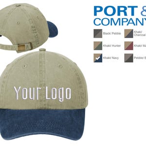 May include: A khaki and navy blue baseball cap with a two-tone design. The front of the cap is khaki and the back is navy blue. The cap has a curved brim and an adjustable strap. The words "Your Logo" are embroidered on the front of the cap.