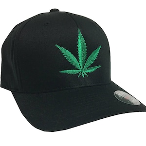 May include: Black baseball cap featuring a green embroidered cannabis leaf design. The cap has a curved brim and a structured fit. A small, round button is on top of the cap.
