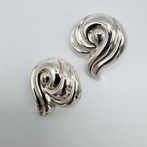 May include: A pair of polished silver-tone earrings featuring a swirling, teardrop-inspired design with smooth, reflective surfaces. The earrings have a rounded, organic shape that tapers to a point, creating a continuous, flowing pattern.