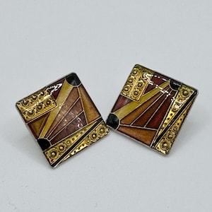 May include: A pair of square-shaped earrings featuring a geometric design with enamel in shades of brown, yellow, and black. The metal edges are gold-toned with a textured, embossed pattern along the sides, creating a distinct, angular appearance.
