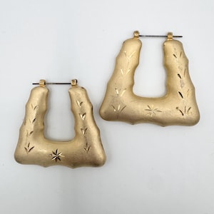 May include: A pair of gold-toned hoop earrings with a square-shaped bamboo-inspired design. The surface features a matte finish with etched leaf-like patterns and a small starburst engraving on the bottom edge of each earring with a metal post.