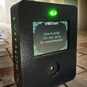 May include: Black rectangular device with a screen displaying the text "SPIRITCHAT" and German text. It has a toggle switch, a green light, and a circular lens. The device has green accents and a textured surface.