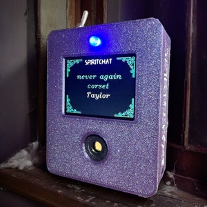 May include: A purple, glittery SpiritChat device with a screen displaying the text "never again corset Taylor." The device has a blue light on top and a circular lens at the bottom. The device is sitting on a wooden surface.