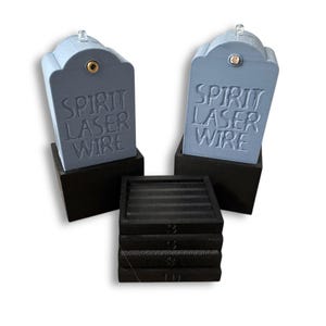 May include: Two light blue, tombstone-shaped objects with the text "SPIRIT LASER WIRE" embossed on the front, each mounted on a black square base. A stack of four black square coasters with horizontal ridges sits in front of the two tombstone objects.