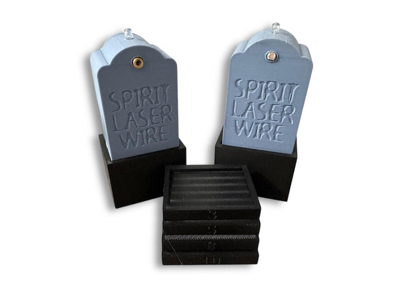 May include: Two light blue, tombstone-shaped objects with the text "SPIRIT LASER WIRE" embossed on the front, each mounted on a black square base. A stack of four black square coasters with horizontal ridges sits in front of the two tombstone objects.