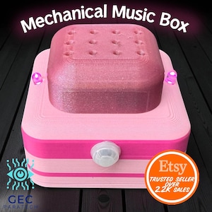 Puede incluir: Una caja de música mecánica rosa con el texto "Mechanical Music Box" en la parte superior. La caja tiene una parte superior cuadrada con varios agujeros y una base rosa claro. El logotipo de Etsy es visible.