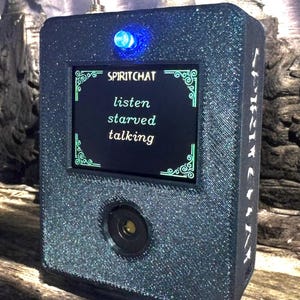 May include: A black, glittery, rectangular device with a blue light and a small screen displaying the text "SPIRITCHAT", "listen", "starved", and "talking".