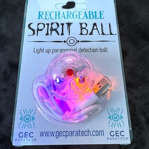 Op de afbeelding: Een oplaadbare spirit ball verpakt op een lichtblauwe kaart. De heldere, bolvormige bal heeft een rood midden en licht op met paarse, blauwe en oranje tinten. De verpakking leest "RECHARGEABLE SPIRIT BALL" en "Light up paranormal detection ball."