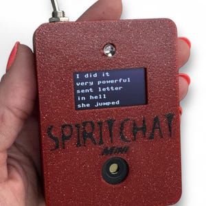 May include: A rectangular red glittery electronic device labeled SPIRIT CHAT MINI with a toggle switch on top, a small screen displaying the text I did it very powerful sent letter in hell she jumped, and a circular button below the text.