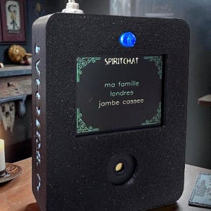 May include: A black rectangular device with the text "SPIRITCHAT" and a screen displaying French text. It has a silver toggle switch on top and a blue light. The device has a lens and decorative accents.