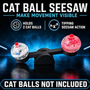Puede incluir: Un juguete de balancín negro para gatos con dos soportes circulares en los extremos. Un soporte contiene una bola texturizada transparente y el otro una bola texturizada roja. Texto: CAT BALL SEESAW, MAKE MOVEMENT VISIBLE, HOLDS 2 CAT BALLS, TIPPING SEESAW ACTION, CAT BALLS NOT INCLUDED.