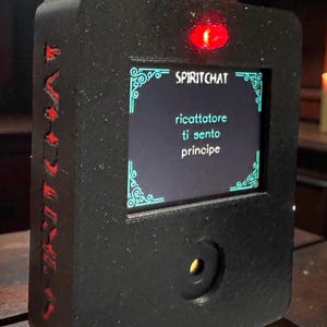 May include: A black rectangular device with the text "SPIRITCHAT" and Italian text on a screen. The device has a red light, a silver toggle switch, and red lettering on the side. The device appears to be a communication tool.